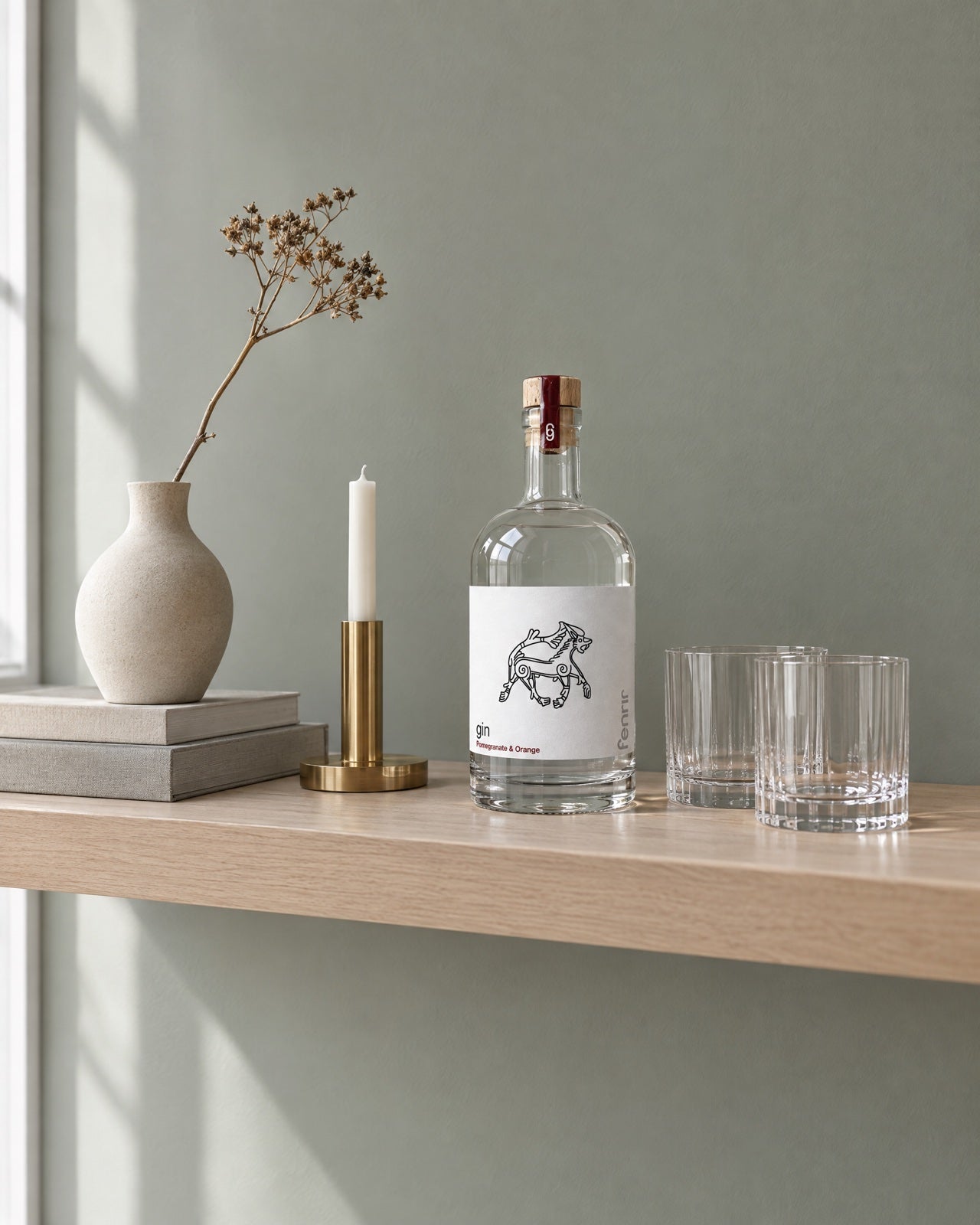 A bottle of Fenrir gin on a pale oak shelf with a vase, a brass candleholder and two tumblers
