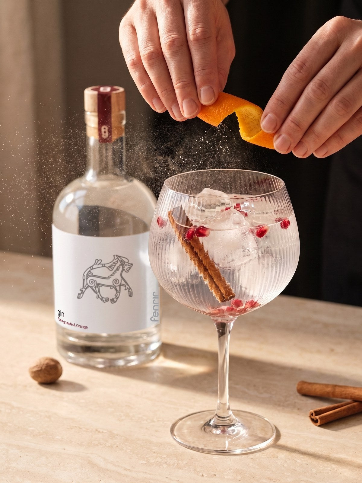 Hands twisting orange peel over a gin and tonic with cinnamon and pomegranate, a bottle of Fenrir behind