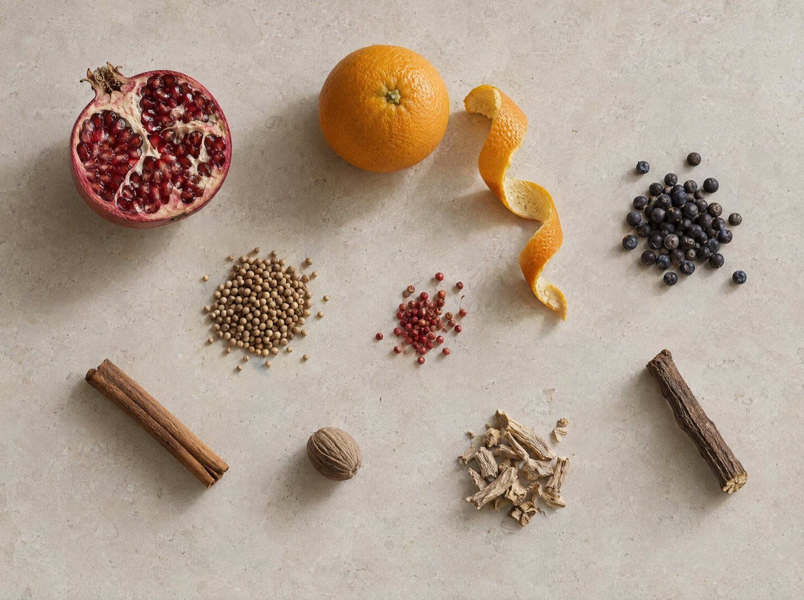 The nine botanicals laid out on pale stone: pomegranate, orange, juniper, coriander, pink pepper, cinnamon, nutmeg, angelica and liquorice