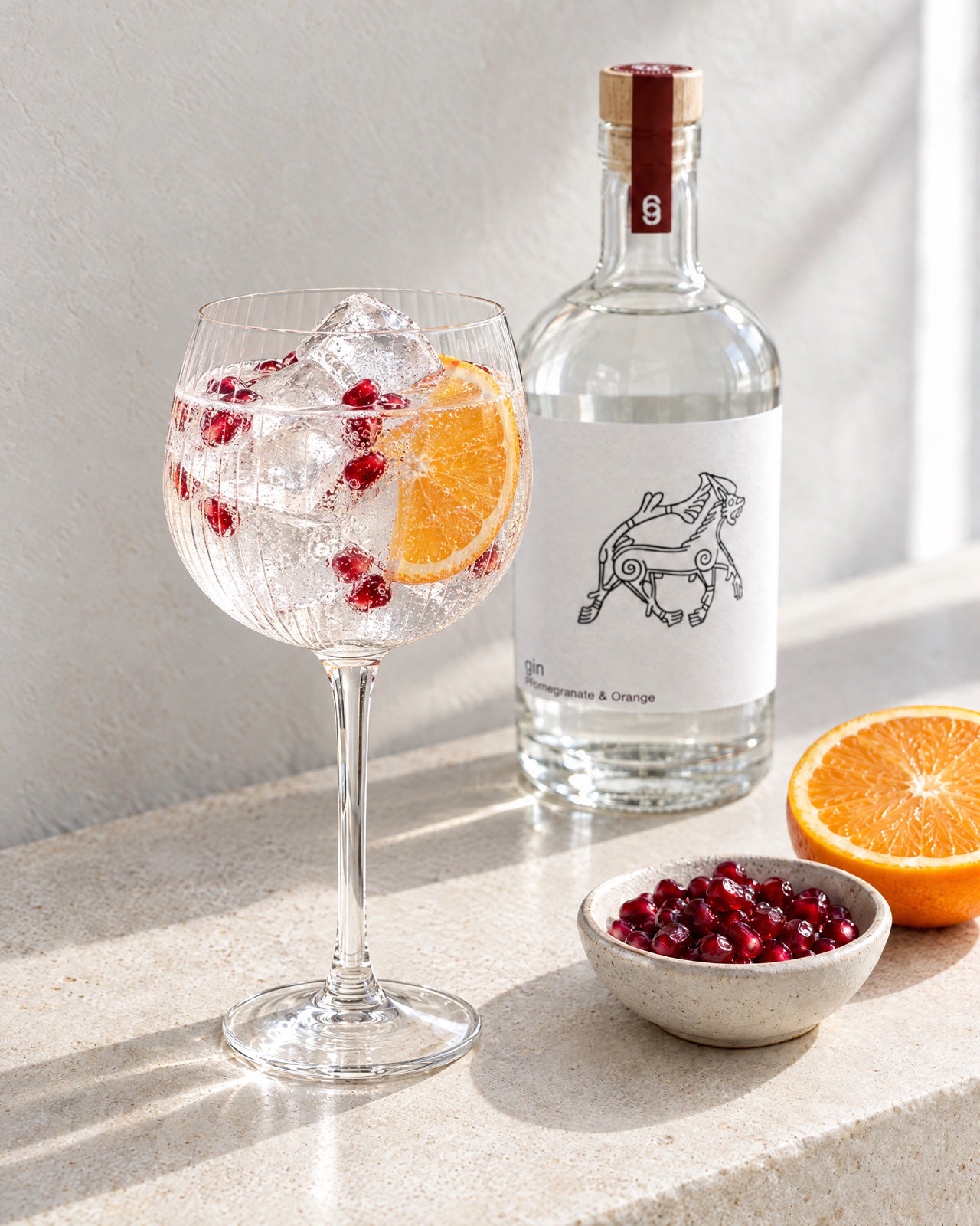A Fenrir gin and tonic in a ribbed balloon glass with orange and pomegranate seeds, on a limestone ledge in morning light