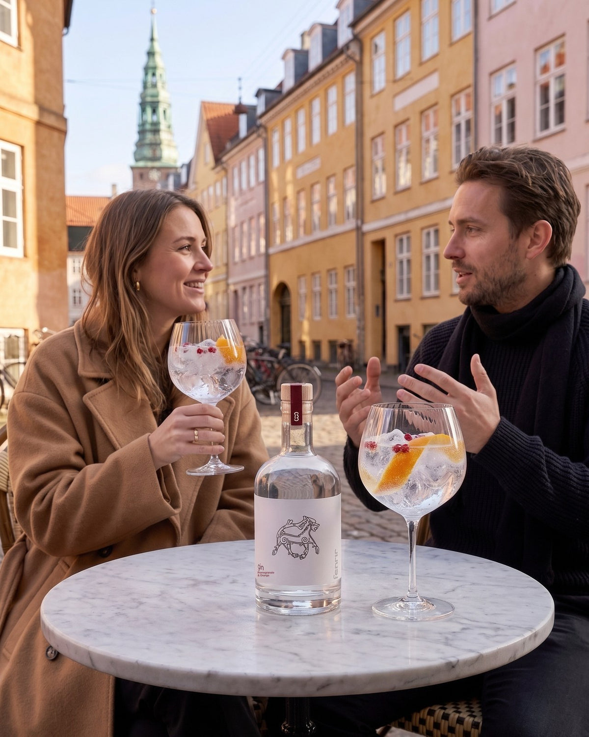 Two friends at an outdoor café table on a cobbled street in Copenhagen with a bottle of Fenrir gin and two gin and tonics
