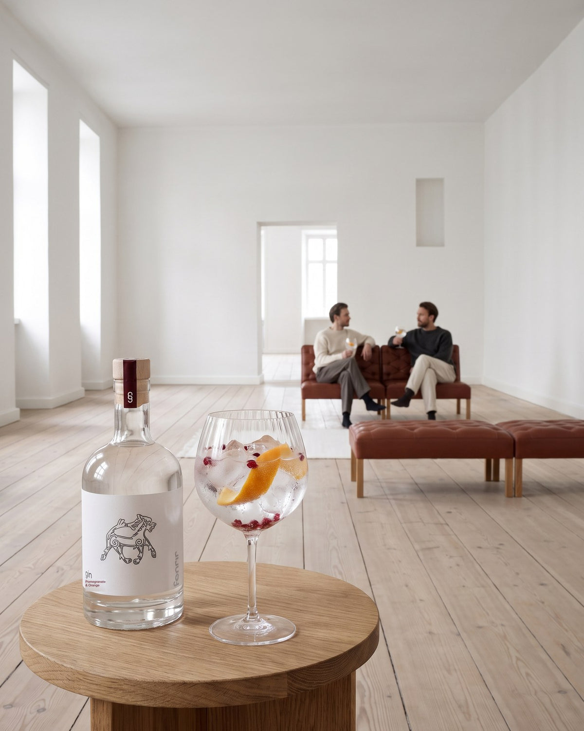 Fenrir gin and a gin and tonic on an oak side table in a bright Copenhagen apartment, two friends on a leather sofa behind
