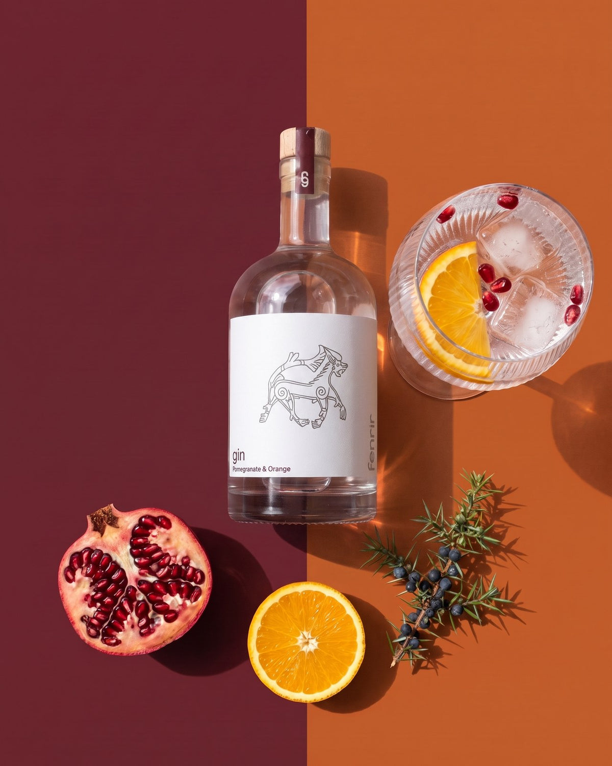 Fenrir gin og en gin og tonic set oppefra på bordeaux og orange, med granatæble, appelsin og enebær
